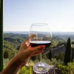 Woman holding a glass of red wine with beautiful landscape of Italy in a background on a sunny day. View from the window.