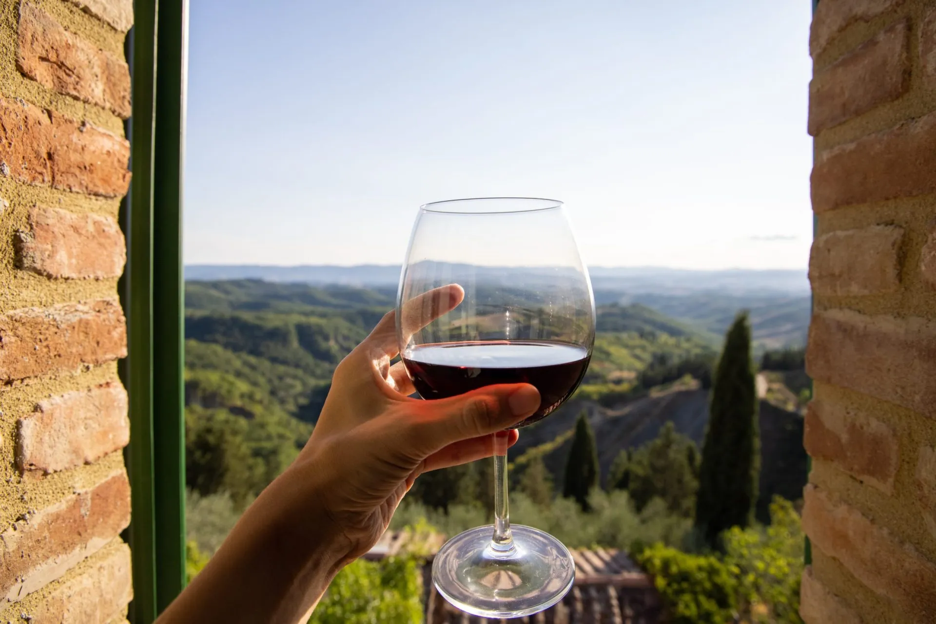 Woman holding a glass of red wine with beautiful landscape of Italy in a background on a sunny day. View from the window.